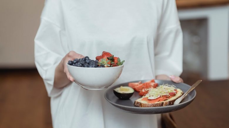 Person holding a bowl of fresh berries and a plate with avocado toast, displaying a healthy breakfast spread.