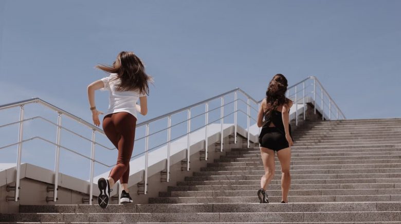 Two women running up outdoor stairs for fitness under a clear blue sky.